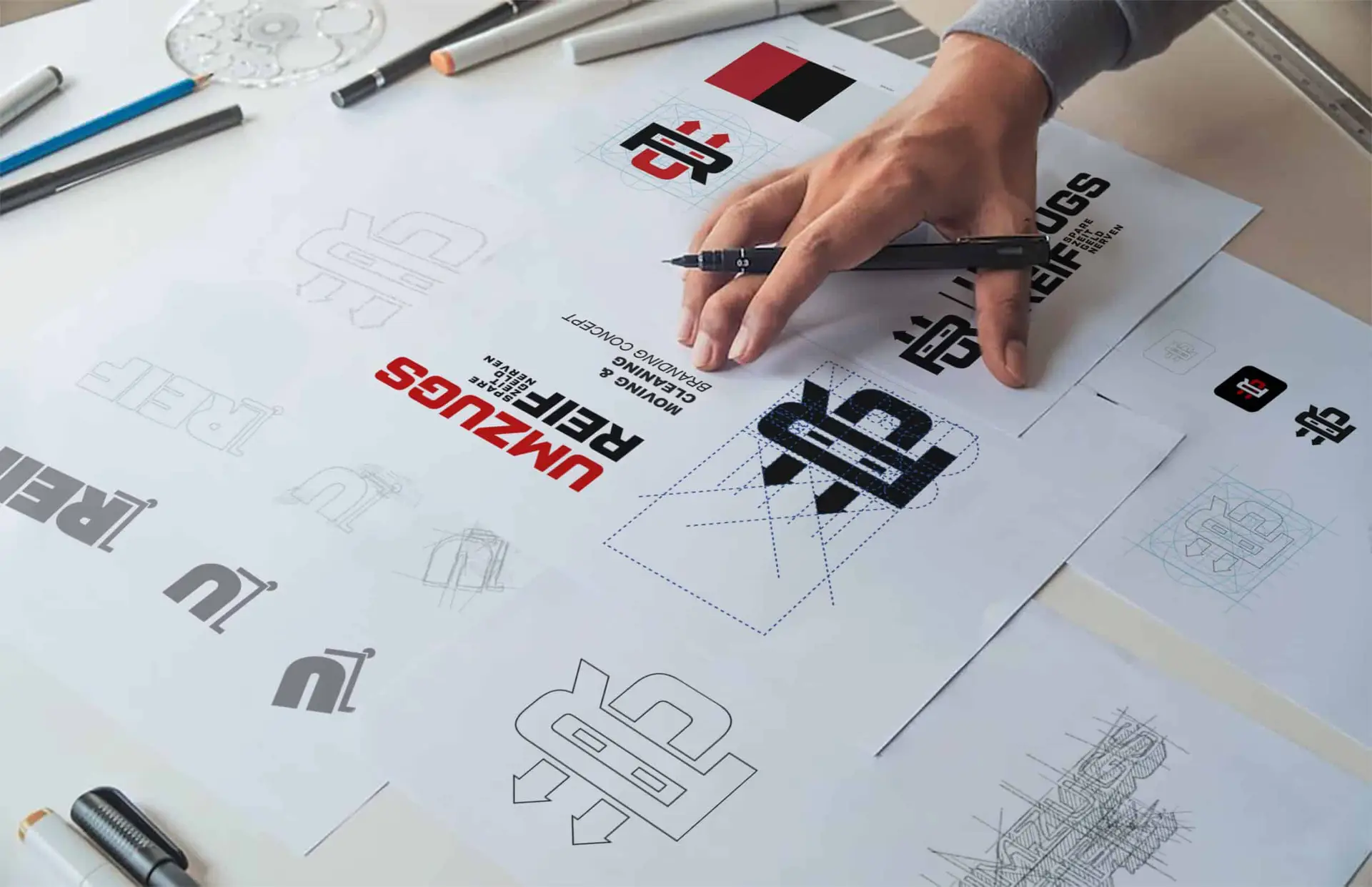Einblick in den kreativen Designprozess von GoldenWing mit Fokus auf die Entstehung eines Firmenlogo, angefangen bei ersten Skizzen bis hin zu finalisierten Versionen in Schwarz und Rot, präsentiert auf einem Arbeitsplatz mit Designwerkzeugen. agentur für branding & webdesign
